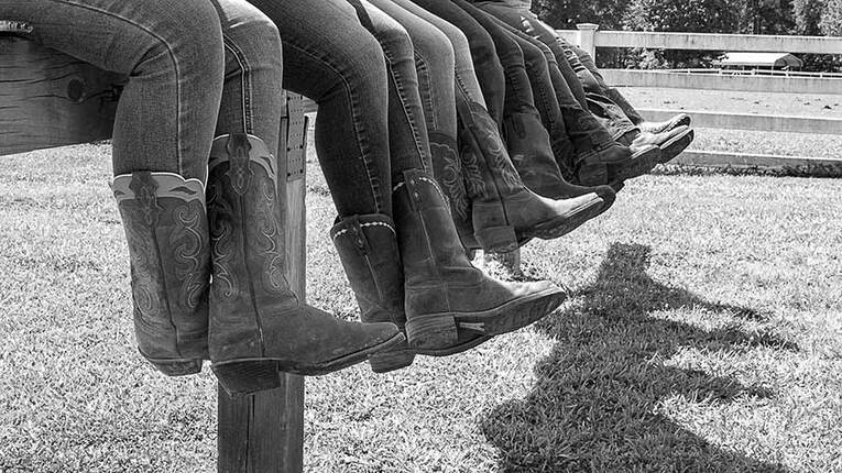 Black and white photo of HOPE board and auxiliary board members' boots in a row, as seen while the members are sitting together on a hitching post. Photo by Jess Schaer.